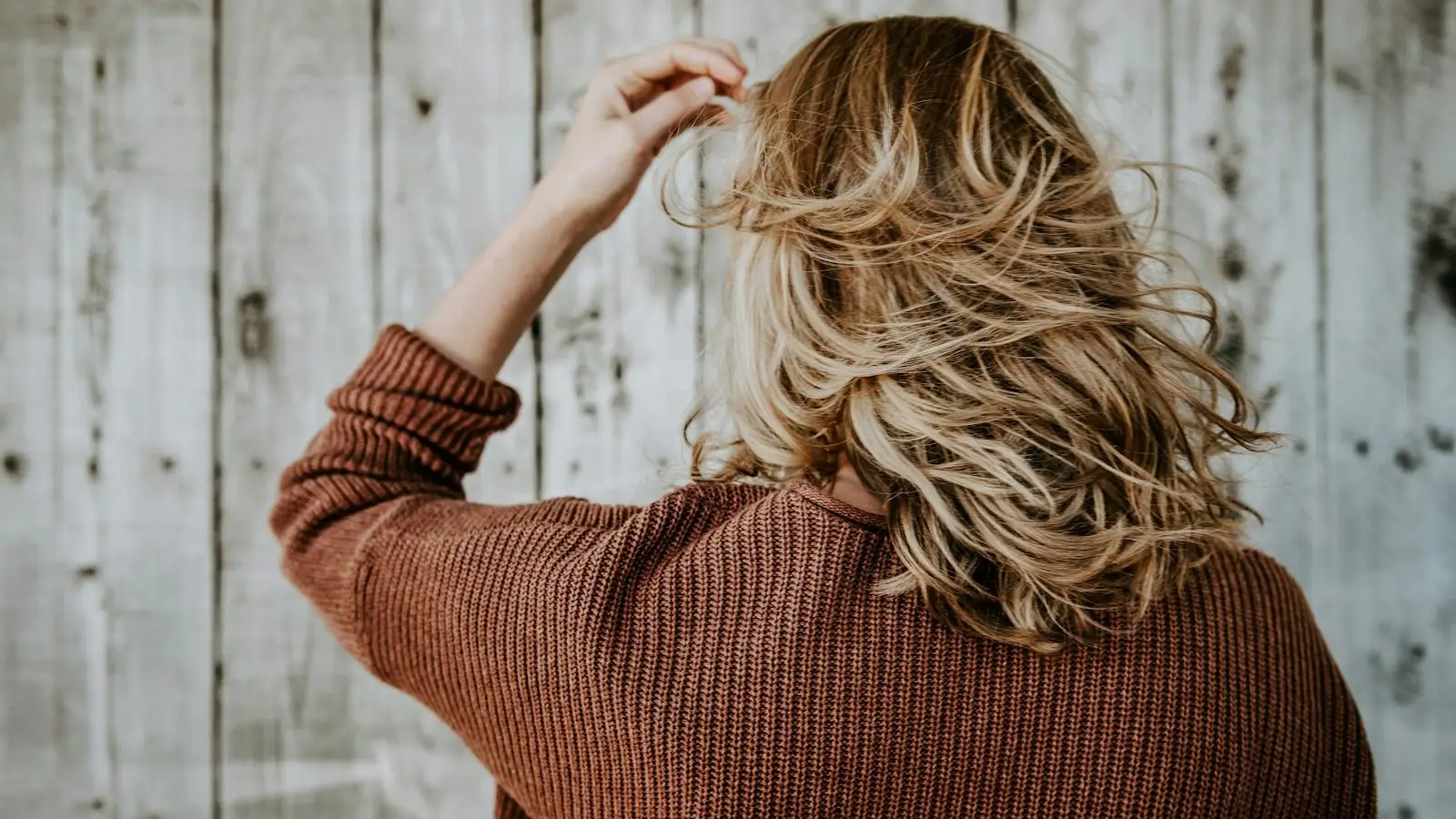 A woman seen from behind touching her shoulder length blonde hair against a pale wooden wall