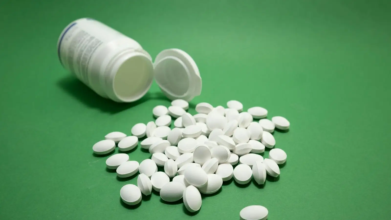 Plain white tablets spilling from an unlabelled white bottle on a green background