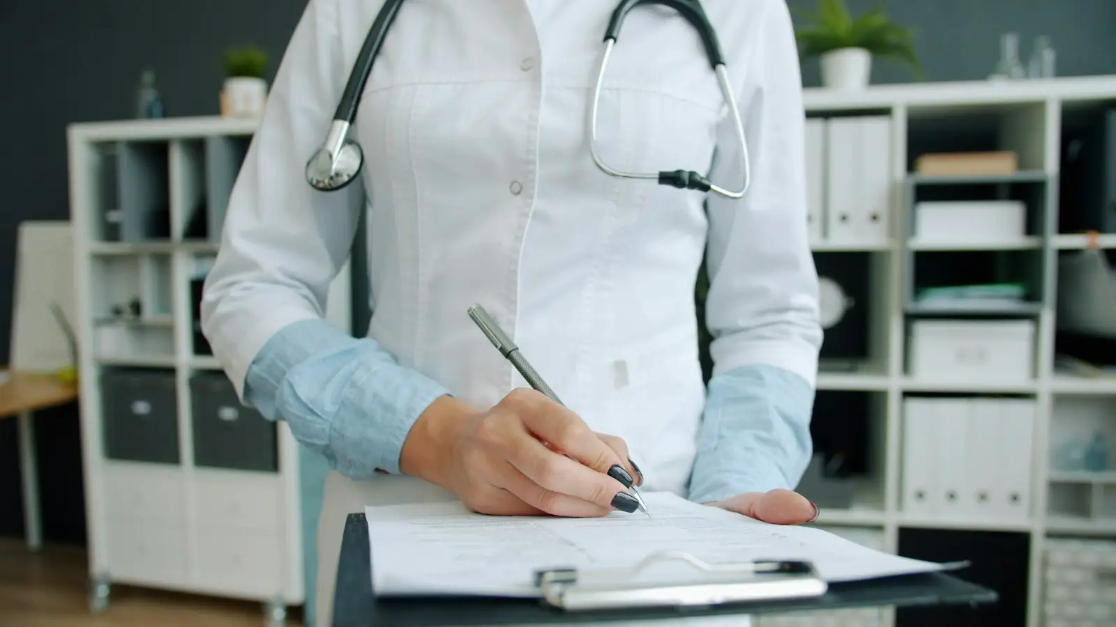A doctor's hands writing on a clipboard in a consulting room
